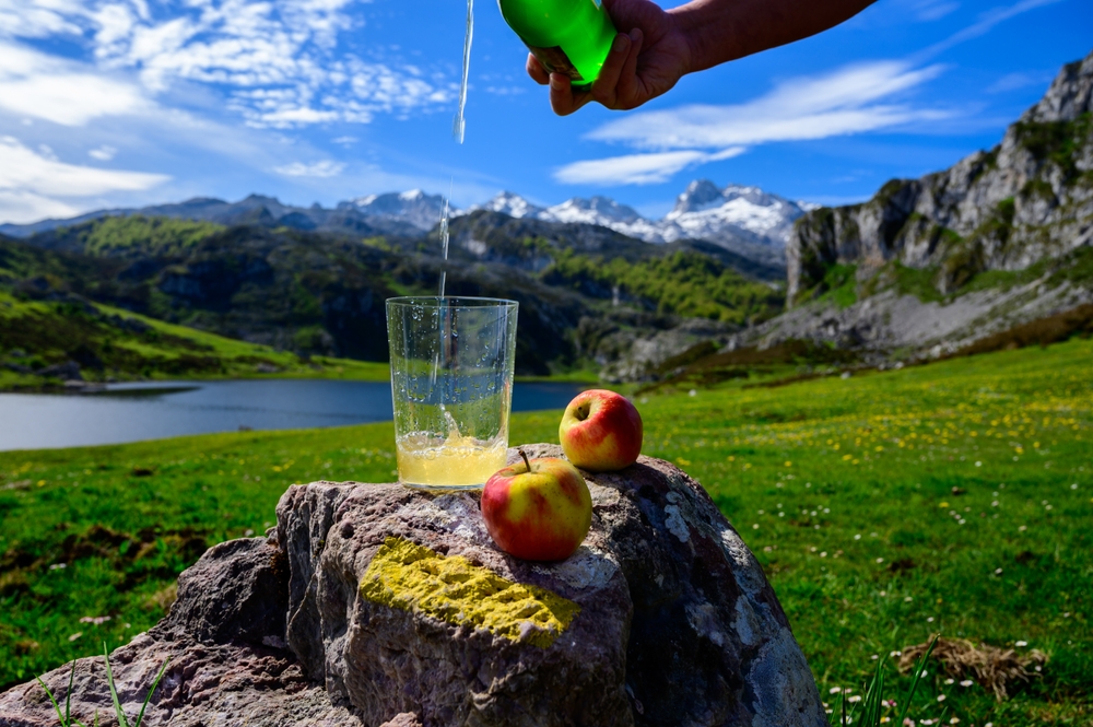 Einschenken von asturischem Apfelwein aus grünen Flaschen aus großer Höhe, hergestellt aus fermentierten Äpfeln, mit Blick auf den Covadonga-See und die Gipfel der Picos de Europa Einschenken von asturischem Apfelwein aus grünen Flaschen aus großer Höhe, hergestellt aus fermentierten Äpfeln, mit Blick auf den Covadonga-See und die Gipfel der Picos de Europa