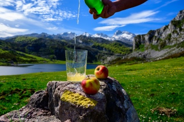 Einschenken von asturischem Apfelwein aus grünen Flaschen aus großer Höhe, hergestellt aus fermentierten Äpfeln, mit Blick auf den Covadonga-See und die Gipfel der Picos de Europa